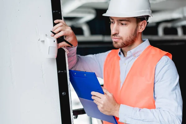 Handsome fireman in helmet checking fire alarm while holding clipboard — Stock Photo