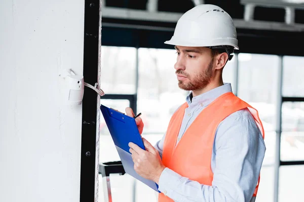 Thoughtful fireman in helmet checking fire alarm while holding clipboard — Stock Photo