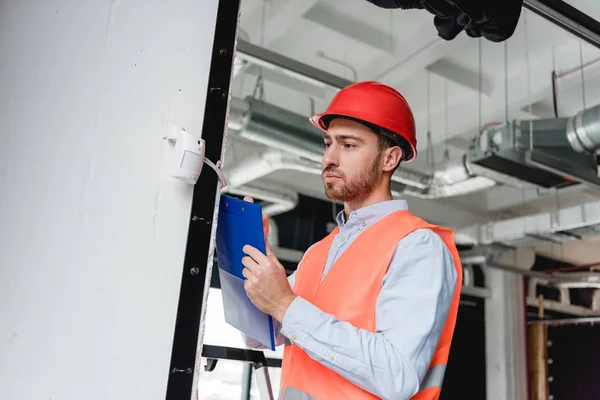 Confident fireman in helmet checking fire alarm while holding clipboard — Stock Photo