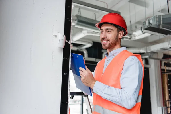 Cheerful fireman in helmet checking fire alarm while holding clipboard — Stock Photo