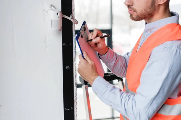 Cropped view of fireman standing near fire alarm while holding clipboard and pen — Stock Photo