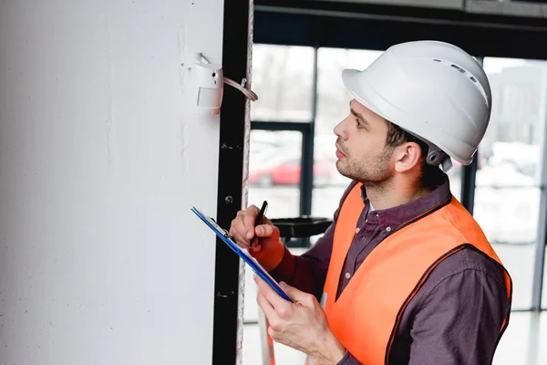 Confident fireman in helmet looking at fire alarm while holding clipboard — Stock Photo