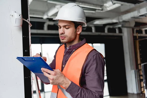 Handsome fireman in helmet standing near fire alarm while looking at clipboard — Stock Photo