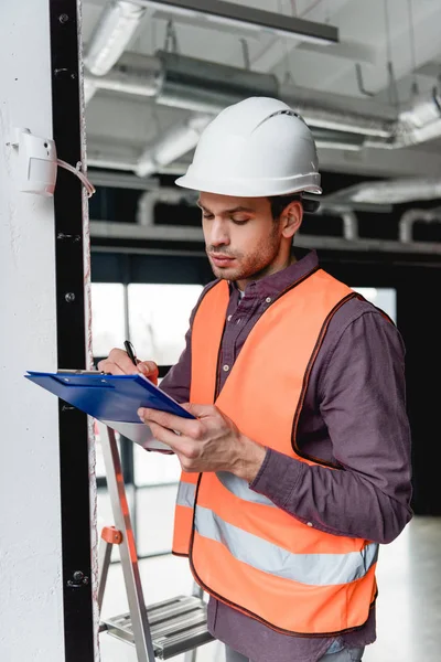 Fireman in helmet standing near fire alarm while writing on clipboard — Stock Photo