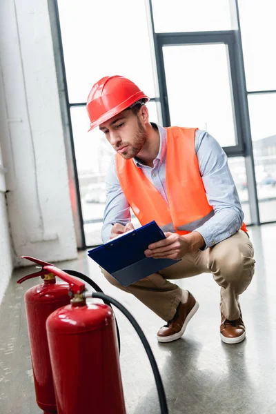 Handsome fireman in helmet checking extinguishers while sitting with clipboard — Stock Photo
