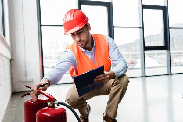 Serious fireman in helmet checking extinguishers while sitting with clipboard — Stock Photo