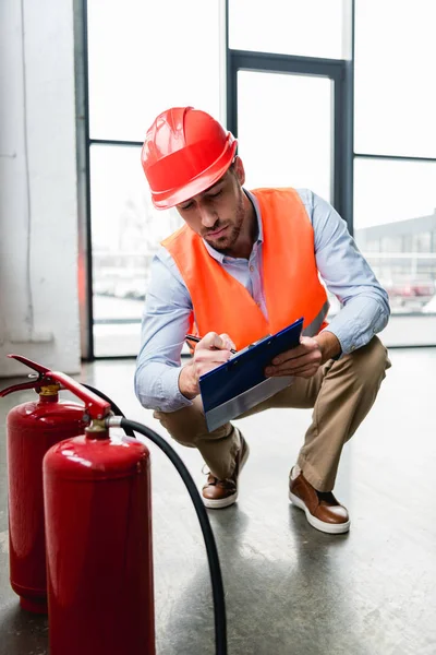 Serious fireman in helmet writing on clipboard while checking extinguishers — Stock Photo