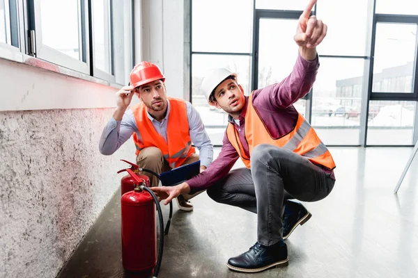 Handsome fireman pointing with finger near coworker while sitting near red extinguishers — Stock Photo