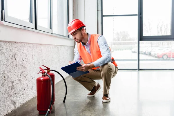 Serious fireman in helmet holding clipboard while looking at extinguishers — Stock Photo