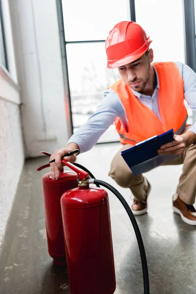 Foco seletivo de extintores vermelhos com bombeiro sério no capacete em segundo plano — Fotografia de Stock