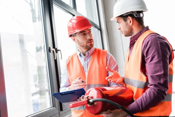 Fireman in helmet looking at coworker while talking and holding red extinguisher — Stock Photo