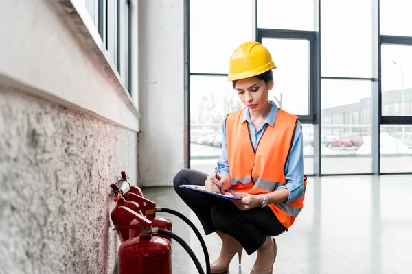 Beautiful firefighter in helmet writing on clipboard while checking extinguishers — Stock Photo