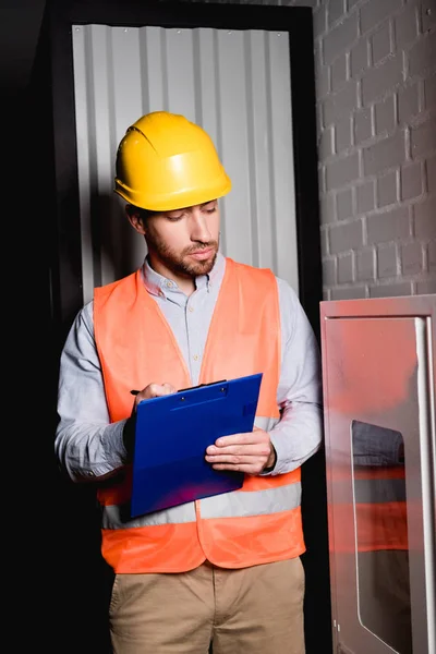 Guapo bombero mirando el panel de fuego mientras sostiene el portapapeles y la pluma — Stock Photo