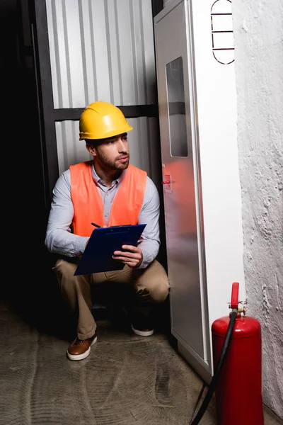 Handsome fireman looking at fire panel while sitting and holding clipboard — Stock Photo