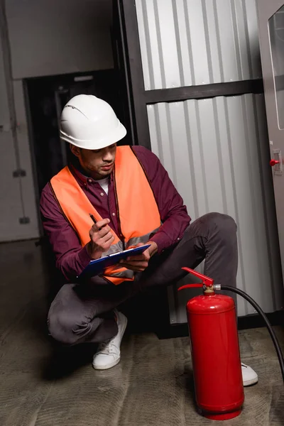 Thoughtful firefighter in helmet sitting and looking at extinguisher near fire panel — Stock Photo