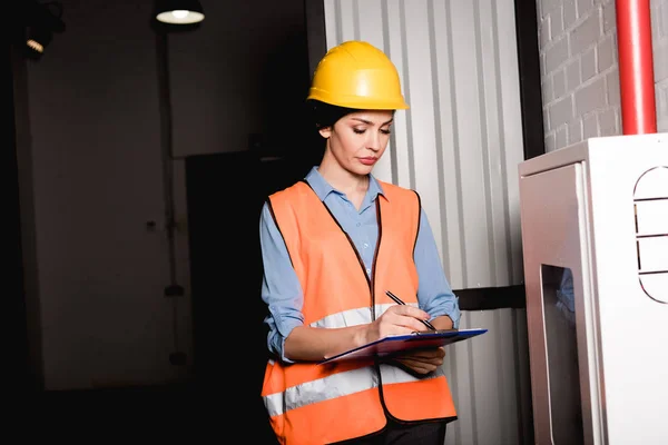 Attractive firefighter in helmet standing near fire panel and writing on clipboard — Stock Photo
