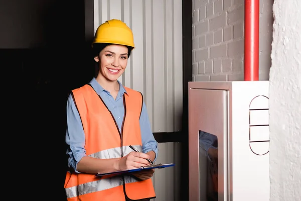 Cheerful female firefighter standing near fire panel and writing on clipboard — Stock Photo