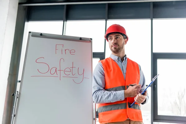 Handsome firefighter in helmet holding clipboard and pen while standing near white board with fire safety lettering — Stock Photo