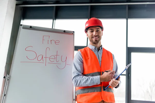 Cheerful firefighter in helmet holding clipboard and pen while standing near white board with fire safety lettering — Stock Photo