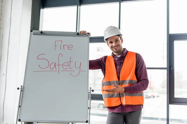 Fröhlicher Feuerwehrmann mit Helm gestikuliert, während er neben Whiteboard mit Brandschutzschriftzug steht — Stockfoto