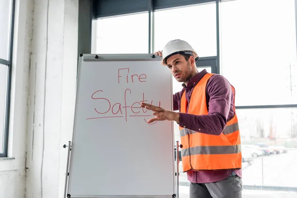 Beau pompier en casque parlant tout en se tenant près du tableau blanc avec lettrage de sécurité incendie — Photo de stock