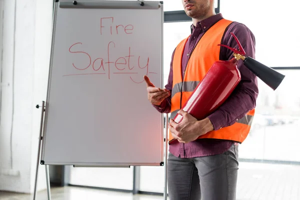 Ausgeschnittene Ansicht eines Feuerwehrmannes, der einen roten Feuerlöscher in der Hand hält, während er neben einer weißen Tafel mit Brandschutz-Schriftzug steht — Stockfoto