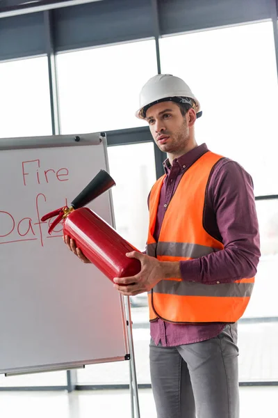 Красивый пожарный держит красный огнетушитель и стоит рядом с белой доской с надписью пожарной безопасности — стоковое фото