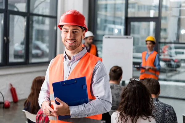 Cheerful fireman holding clipboard near coworkers giving talk on briefing — Stock Photo