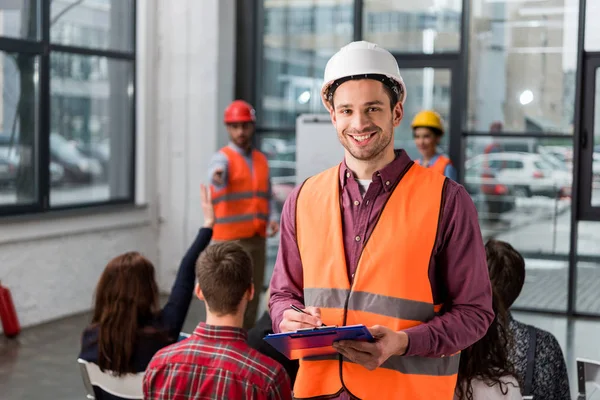 Selektiver Fokus eines glücklichen Feuerwehrmannes mit Klemmbrett in der Nähe eines Mitarbeiters, der einen Vortrag über Einweisung hält — Stockfoto