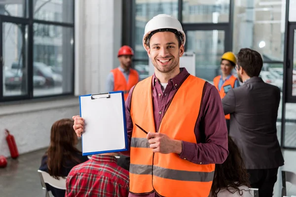 Selektiver Fokus des gut gelaunten Feuerwehrmannes, der mit Stift auf leeres Klemmbrett zeigt, in der Nähe von Mitarbeitern, die einen Vortrag über Hintergründe halten — Stockfoto