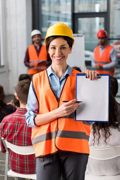 Selektiver Fokus einer fröhlichen Feuerwehrfrau, die mit Stift auf ein leeres Klemmbrett zeigt, in der Nähe von Mitarbeitern, die einen Vortrag über Hintergründe halten — Stockfoto