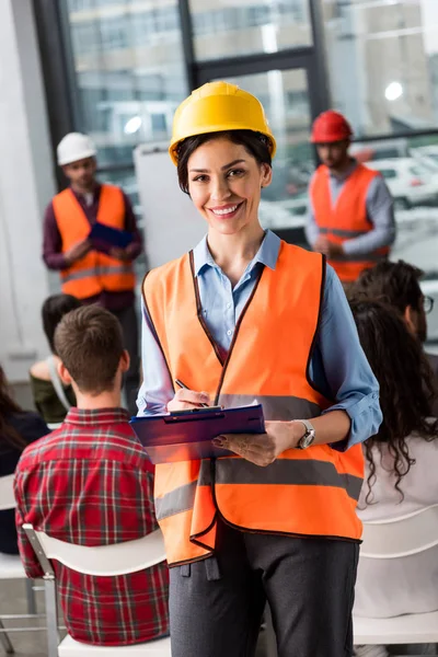 Happy female firefighter holding clipboard near coworkers giving talk on briefing on background — Stock Photo