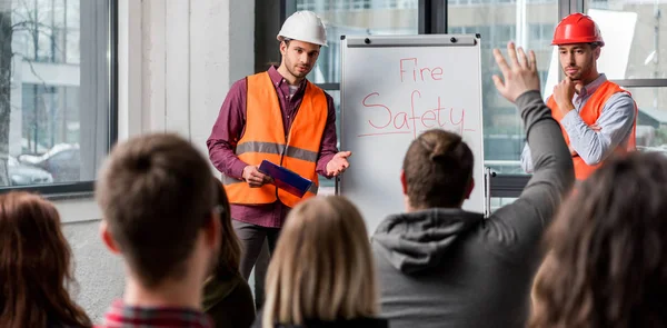 Foco seletivo de bombeiros bonitos em capacetes dando palestra sobre briefing perto da placa branca com letras de segurança contra incêndio — Fotografia de Stock