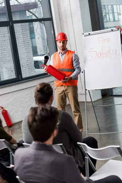 Handsome fireman in helmet giving talk on briefing while holding extinguisher near white board with fire safety lettering — Stock Photo