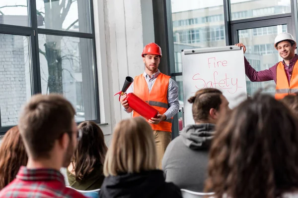 Selective focus of handsome fireman in helmet holding extinguisher while standing near white board with fire safety lettering near coworker — Stock Photo