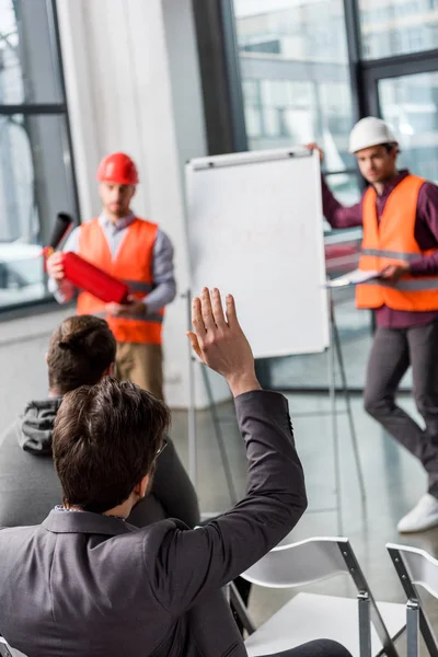 Selective focus of man raising hand near handsome firemen in helmets standing near white board with fire safety lettering — Stock Photo