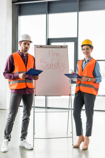 Selective focus of fireman standing near attractive coworker near white board with fire safety lettering — Stock Photo