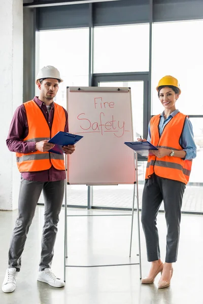 Selektiver Fokus des Feuerwehrmannes steht neben attraktivem Mitarbeiter neben Whiteboard mit Brandschutz-Schriftzug — Stockfoto