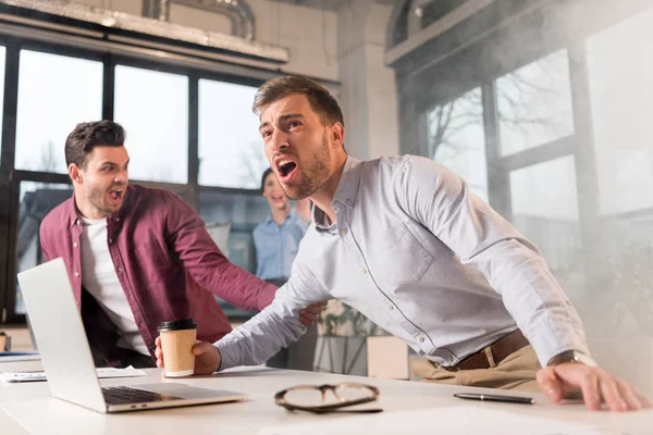 Scared businessman holding paper cup and yelling near colleagues in office with smoke during fire — Stock Photo