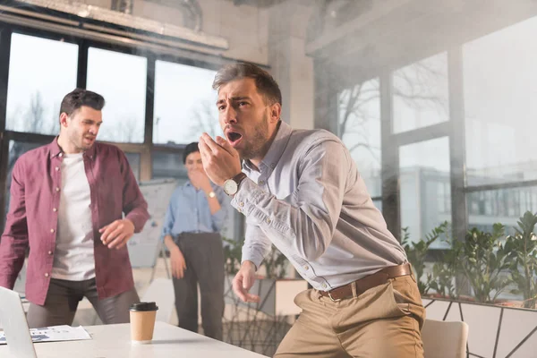 Terrified businessman standing with open mouth near colleagues in office with smoke — Stock Photo