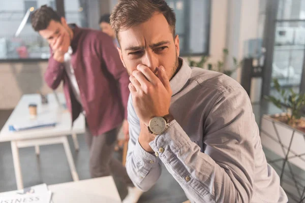 Hombre guapo sosteniendo la nariz cerca de compañeros de trabajo en la oficina con humo - foto de stock