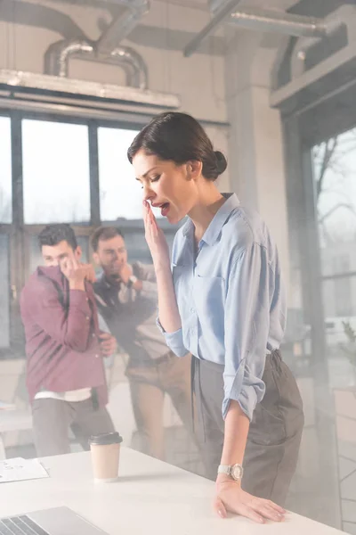 Empresaria de pie con la boca abierta cerca de compañeros de trabajo en la oficina con humo - foto de stock