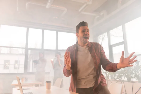 Guapo hombre de negocios gritando en la oficina con humo cerca de compañeros de trabajo - foto de stock