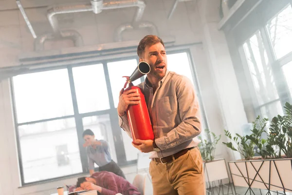 Scared businessman holding extinguisher and screaming in office with smoke near coworkers — Stock Photo