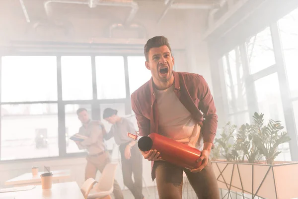 Empresario sosteniendo extintor y gritando en la oficina con humo cerca de compañeros de trabajo - foto de stock