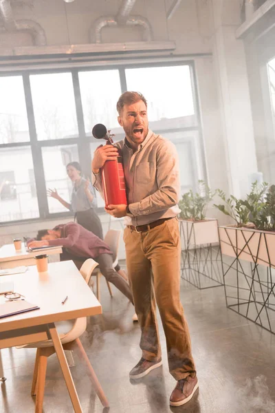 Empresario sosteniendo extintor y gritando en la oficina con humo cerca de compañeros de trabajo - foto de stock