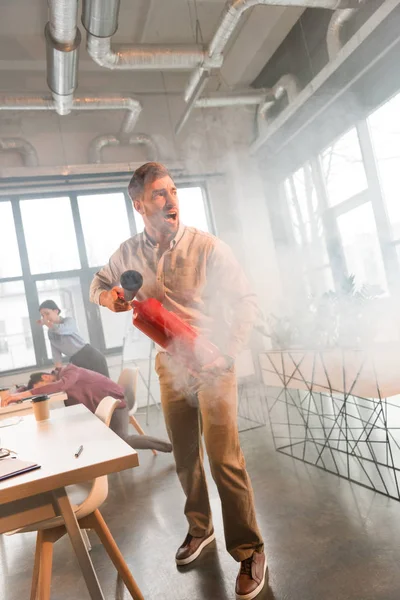 Schöner Geschäftsmann hält Feuerlöscher in der Hand und schreit im Büro mit Rauch in der Nähe von Kollegen — Stockfoto