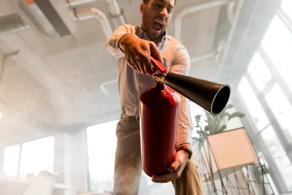 Schöner Geschäftsmann hält Feuerlöscher in der Hand und brüllt im Büro mit Rauch — Stockfoto