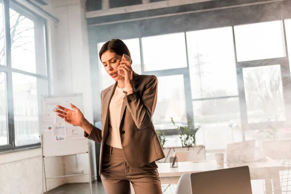 Beautiful scared businesswoman talking on smartphone in office with smoke — Stock Photo