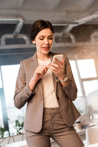 Verängstigte Geschäftsfrau blickt im Büro mit Rauch auf Smartphone — Stockfoto